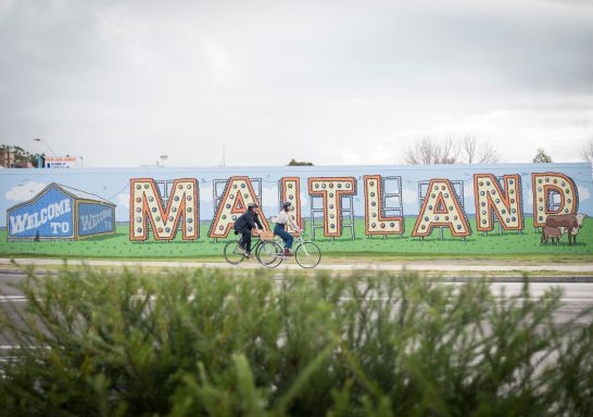 Couple riding their bicycles past the Welcome to Maitland Sign on Elgin St, Maitland